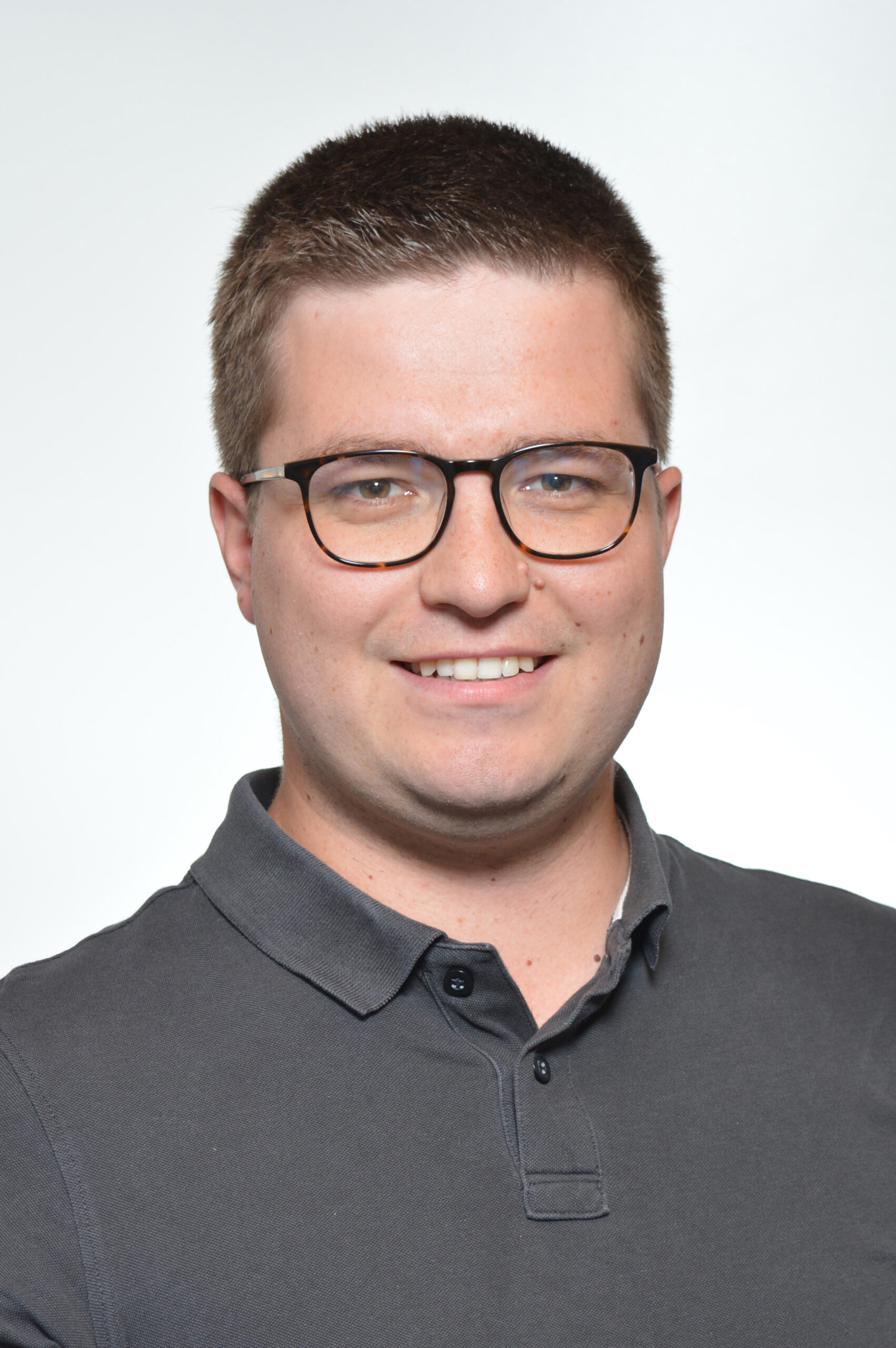 Kandidaten-Profilbild_sehlen_ Porträtaufnahme von Kandidat Sehlen mit Brille und grauem Polo-Shirt, lächelnd vor hellem Hintergrund.